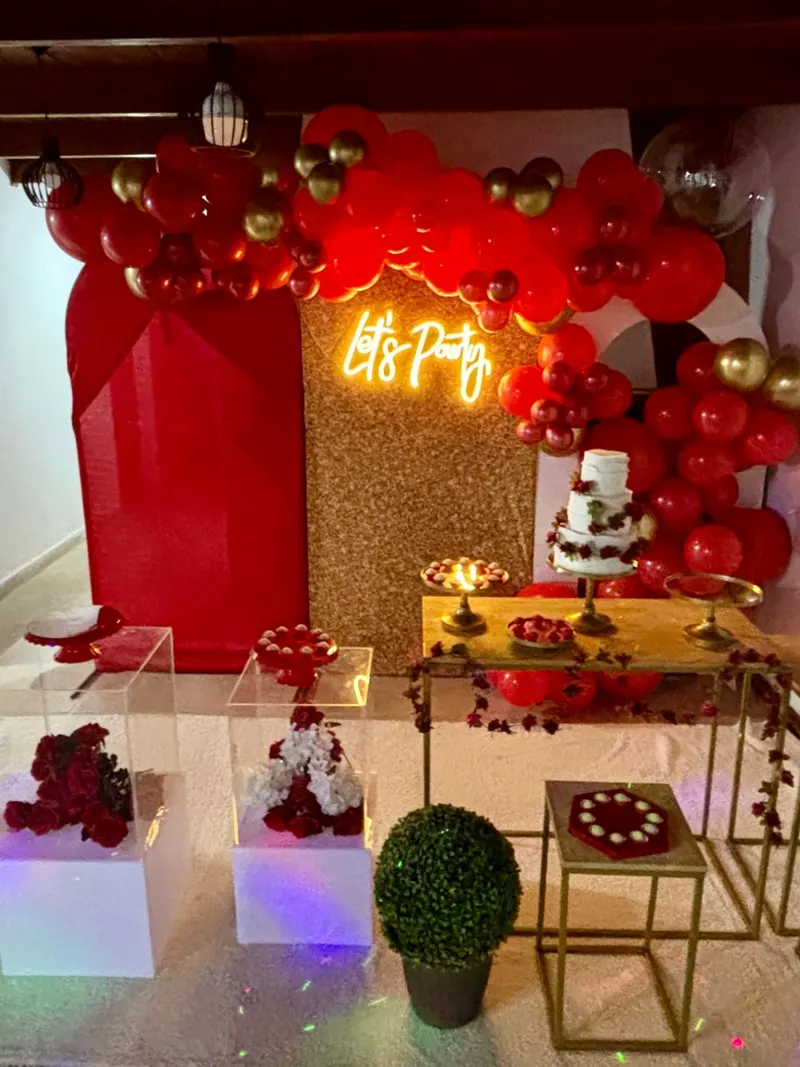 Mesa de festa com arco de balões vermelhos e dourados, painel vermelho e neon “Let’s Party”